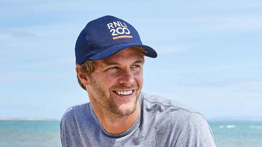 A blonde man wearing a dark blue baseball cap with ‘RNLI 200’ embroidered across the front. He’s at the beach, smiling, with blue sea and sky behind him. 