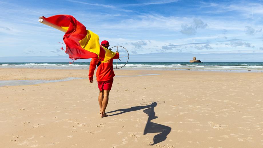 An RNLI lifeguard in a red sweater, cap and shorts, carries a beach flagpole down the beach towards the waterline