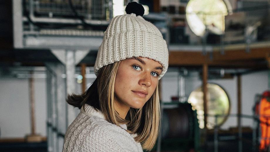 A woman with blonde hair looks at the camera, wearing a cream-coloured beanie with a navy bobble on top
