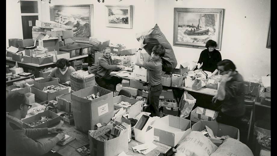 People working at desks sorting through buttons and postcards that were sent in to raise money for the RNLI as part of a Blue Peter appeal. The room is cluttered, with overflowing boxes and sacks, as well as large piles of paperwork.