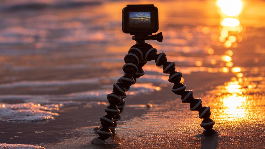 A small camera on a shore-mounted tripod stands on the shoreline. The sun is setting behind it.