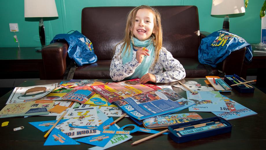 A young girl sits and reads the 30th anniversary edition of Storm Force magazine on a table covered in puzzles, activities and drawing materials