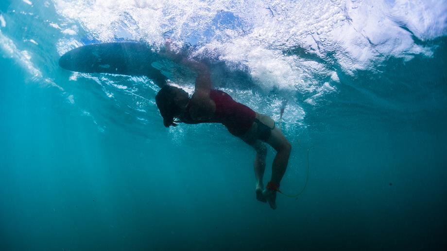 Underwater image of a surfer demonstrating a turtle roll manoeuvre in clear water. The surfboard is upside down above them, while they hold on to the sides.