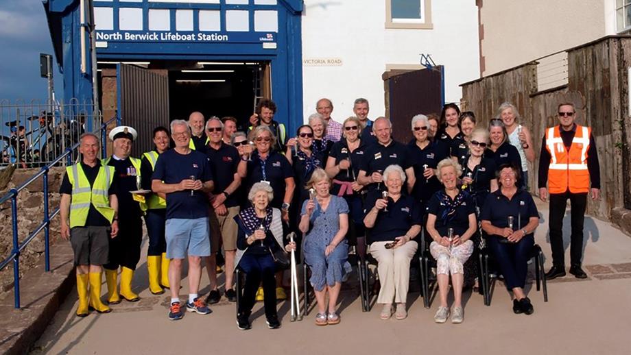 A group shot of 29 RNLI volunteers on the slipway across the road from North Berwick Lifeboat Station. They are smiling and raising a glass to 200 years of saving lives at sea.