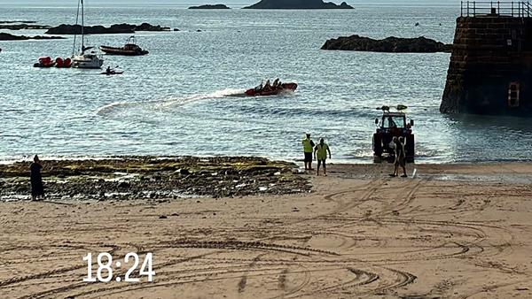 A view across sand and out beyond a harbour wall to the open sea, showing a silhouette of the lifeboat leaving the harbour. The RNLI launching tractor and shore crew are at the water’s edge, seeing them off.
