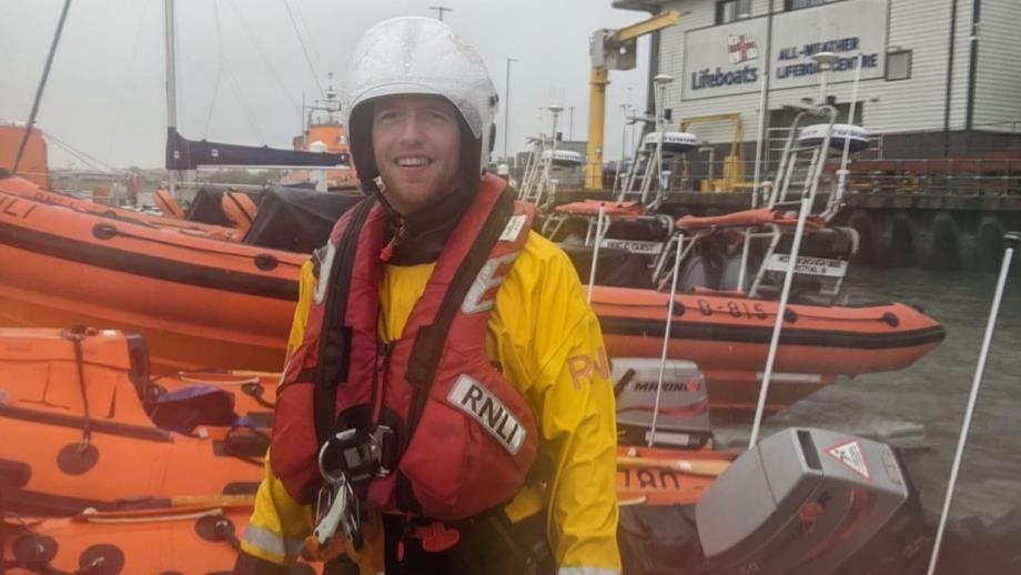 Anstruther volunteer Crew Member and Helm Tam Bett in full crew kit. He is standing outside in front of a row of D class inshore lifeboats at the All-weather Lifeboat Centre in Poole, Dorset.