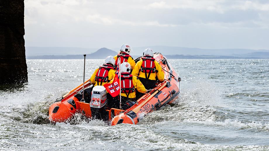 The back of four Anstruther lifeboat volunteers in their D class inflatable lifeboat as it heads out to sea. The sea is calm and it’s a bright but cloudy day.