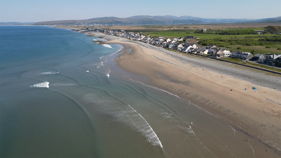Borth Beach – the calm blue sea meets a yellow sandy beach that has houses and green fields behind it, with large hills in the background.