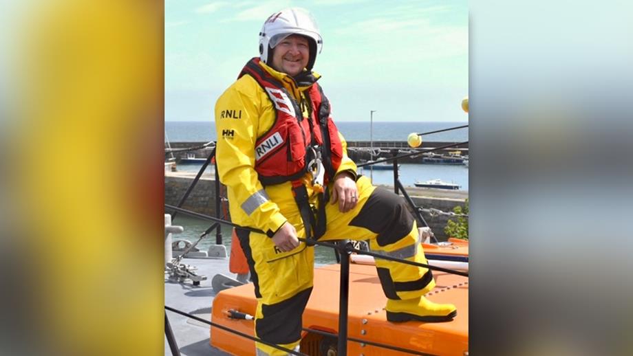 Brendan Rooney, Crew Member at Newcastle RNLI, standing on an all-weather lifeboat in full crew kit. The sky is blue with a few small clouds, and the sea is in the background.