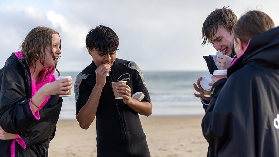 Teenagers talking on the beach with hot drinks, wearing Dryrobes and wetsuit