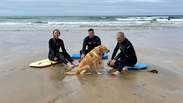 Richard, Charlotte and Ed are sitting on their bodyboards on the sand, wearing wetsuits and still damp from the sea. They’re all smiling and their dog, Flo, sits in the middle of them. 