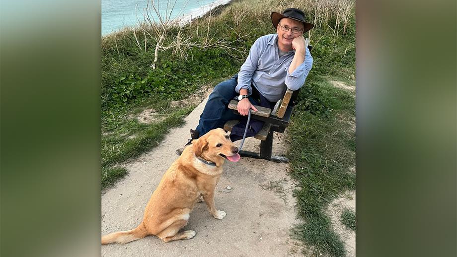 Richard, who is wearing glasses and a leather outback hat, smiles at the camera while sitting on a bench, holding the lead of his dog