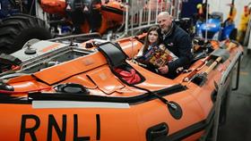 Guy Addington, Margate Crew Member and Regional Water Safety lead, sitting in an inshore lifeboat. His daughter Elodie is sitting in front of him in the lifeboat. Guy is holding a Storm Force magazine in his left hand. Both Guy and Elodie are smiling at the camera. There are other lifeboats and equipment in the background.