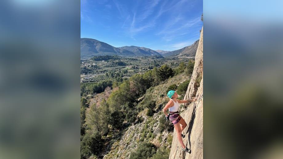 Jo high up on a rock face in Alicante, Spain, using safety ropes and wearing a safety helmet. Below her is a lush green valley surrounded by mountains. It’s a bright, sunny day. 