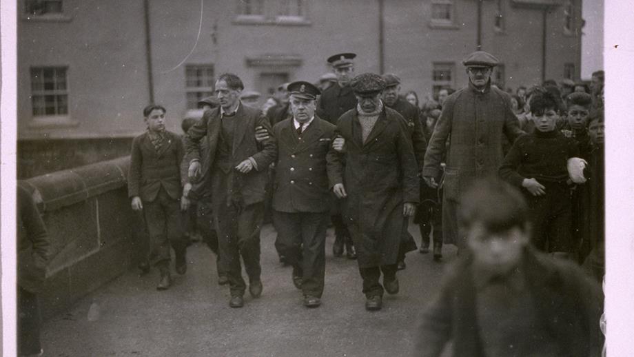 A black and white photograph showing a uniformed coastguard officer holding the arms of two of the crew from the Foremost and helping them walk them down a busy road, which is crowded with people. 