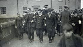 A black and white photograph showing a uniformed coastguard officer holding the arms of two of the crew from the Foremost and helping them walk them down a busy road, which is crowded with people. 