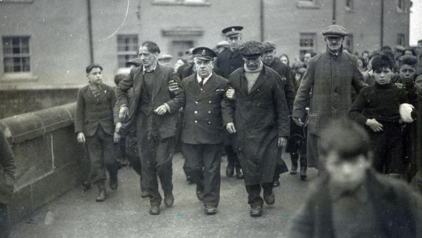 A black and white photograph showing a uniformed coastguard officer holding the arms of two of the crew from the Foremost and helping them walk them down a busy road, which is crowded with people. 
