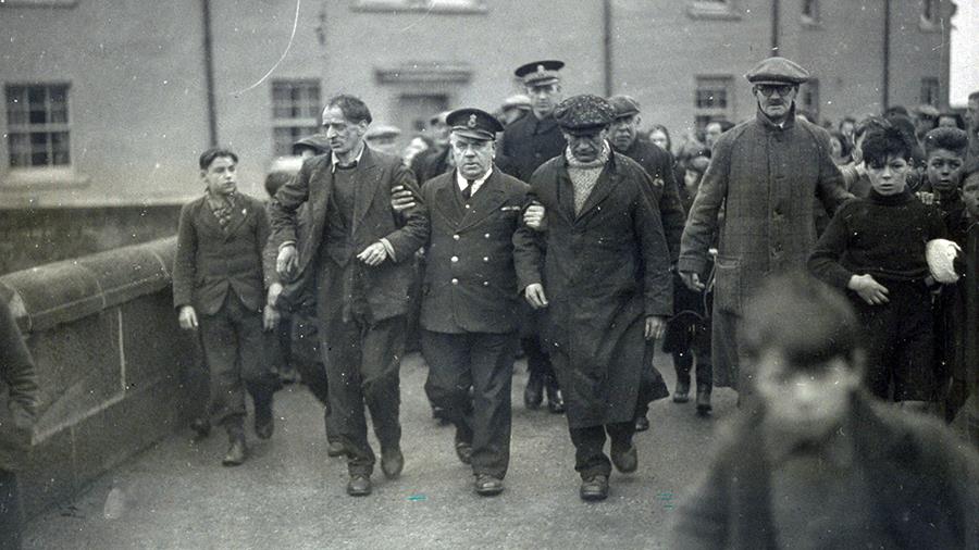 A black and white photograph showing a uniformed coastguard officer holding the arms of two of the crew from the Foremost and helping them walk them down a busy road, which is crowded with people. 