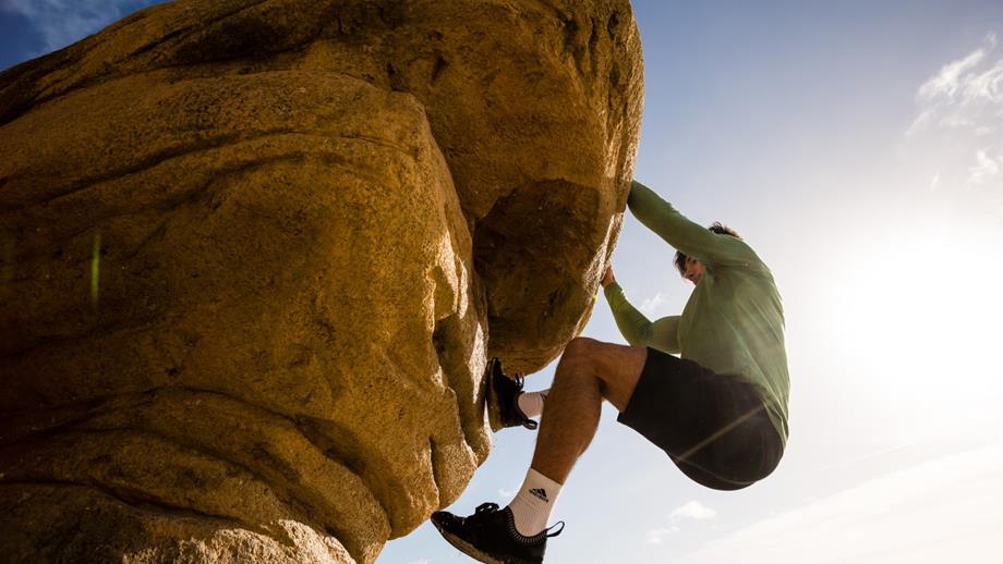A boulderer hanging from a tall rock. He is wearing shorts and a long-sleeved top. It’s a bright, sunny day.