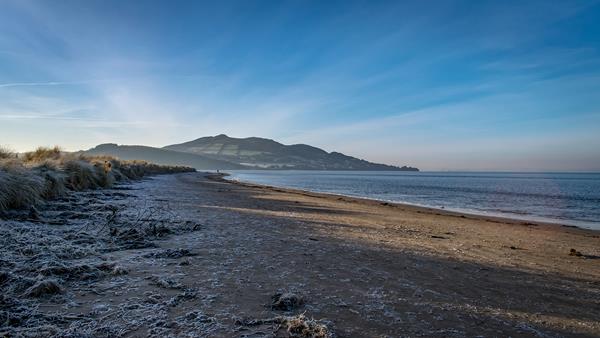 A long sandy beach edged with marshes and covered in frost. Rolling hills with fields and trees can be seen in the distance.