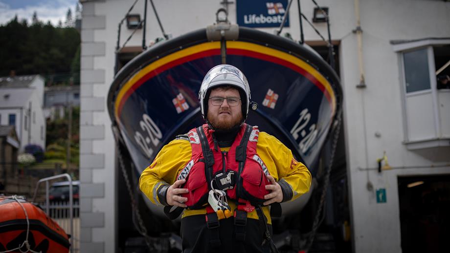 Lochlainn Leneghan, Helm at Newcastle RNLI, standing in front of the station’s all-weather lifeboat. He is wearing crew kit and is holding onto either side of the lifejacket that he is wearing. He is looking at the camera with a neutral expression.