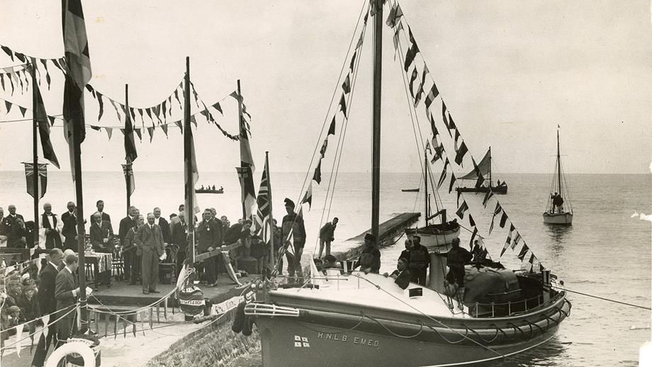 A black and white photograph of the E.M.E.D's naming ceremony in 1930. The lifeboat is moored in a harbour and decked out with bunting. 