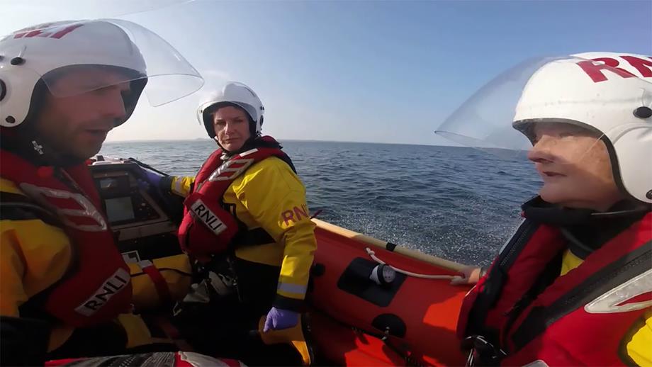 Three crew are afloat on an orange RNLI inshore lifeboat with the open sea in the background. They are making their way to the scene of a rescue so are discussing a plan of action.