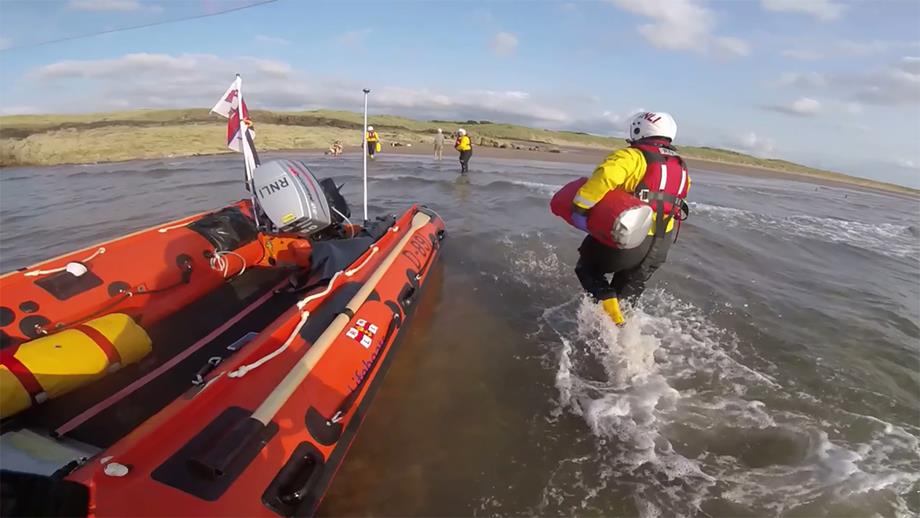 A helmet camera view from near the bow of the inshore lifeboat towards a sandy beach framed by low-lying rocks. Three crew members wade through shallow water towards a casualty who can be seen sitting at the water’s edge.
