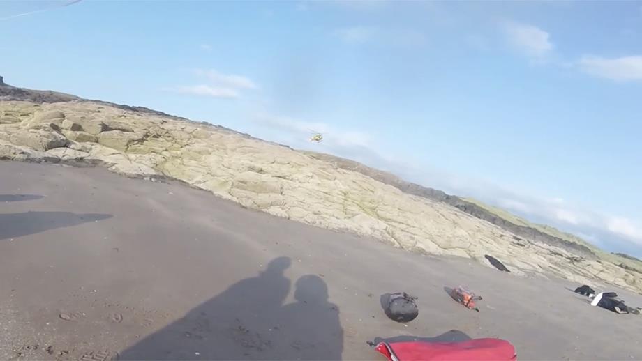 A helmet camera view from a sandy beach up over low-lying rocks. A helicopter can be seen approaching in the distance