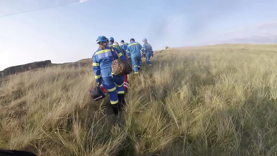 A helmet camera view across long grass of a group of coastguards and helicopter crew transporting the casualty to the helicopter for transfer to the hospital.