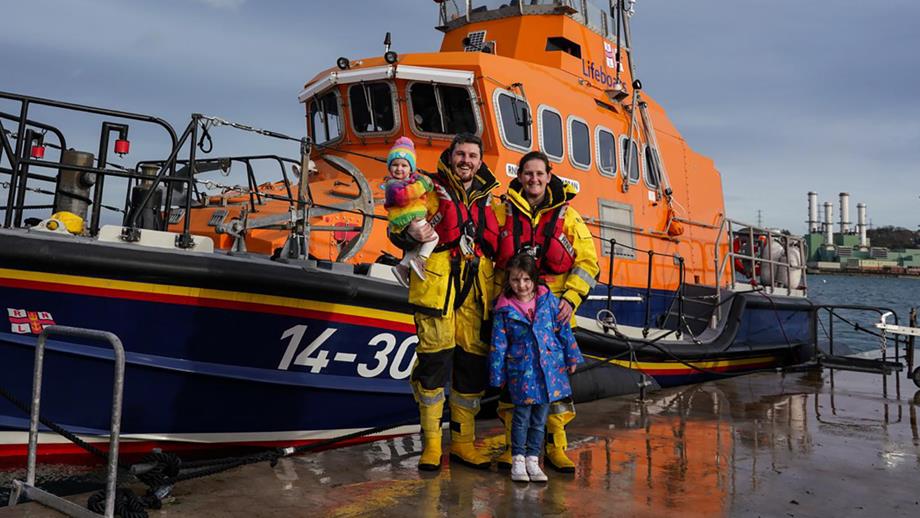 Pamela stands next to her husband Scott, alongside their two young daughters, in front of the Larne RNLI’s Trent class lifeboat. Pamela and Scott are both in all-weather crew kit, and are smiling at the camera.