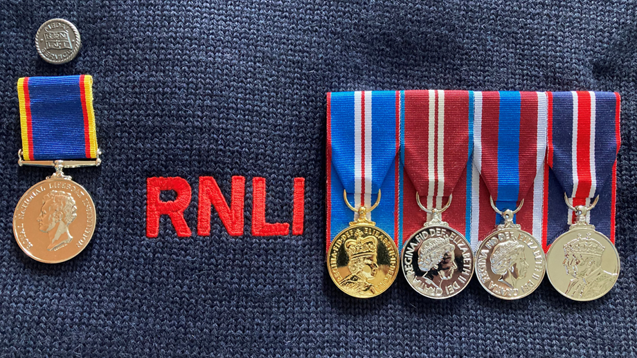 The medals of RNLI Crew Member Dave Riley, pinned to a navy blue Guernsey.  On the left is an RNLI Long Service Medal with a Silver Medal service badge above. On the right is a Golden Jubilee Medal, Diamond Jubilee Medal, Platinum Jubilee Medal and King's Coronation Medal.
