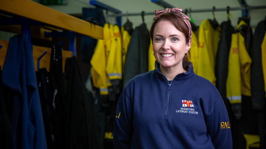 Lexi stands in the changing room at Mudeford Lifeboat Station. You can see crew kit hanging up behind her. Lexi is wearing a navy blue Mudeford Lifeboat Station fleece, a pink and brown striped headband, and gold earrings. She’s smiling at the camera.