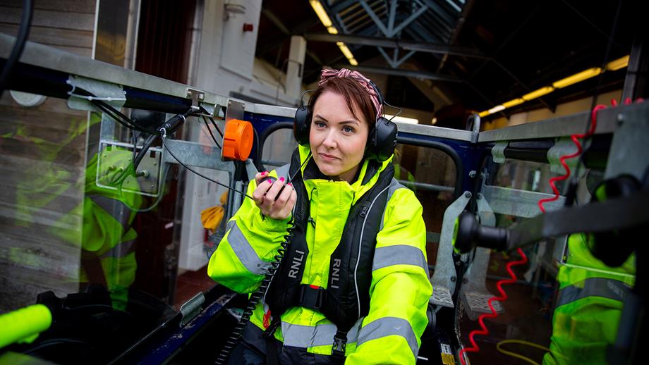 Lexi, wearing a pink headband and a high-vis jacket, is driving the station’s tractor. She’s wearing headphones and talking into a radio. She looks focused and determined. 