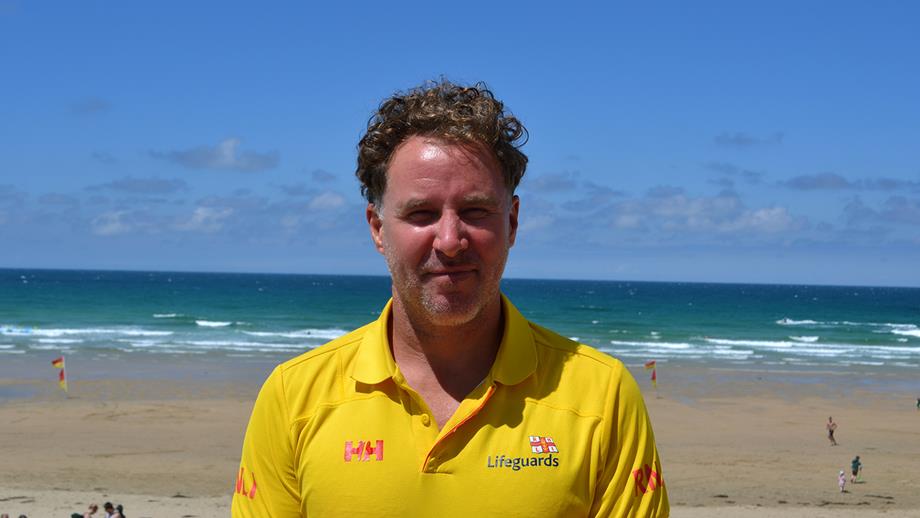 Steve, a man with curly brown hair, smiles at the camera. He’s wearing a yellow lifeguard polo shirt. Behind him is a golden beach with turquoise waters, and a red and yellow flag in the distance. 