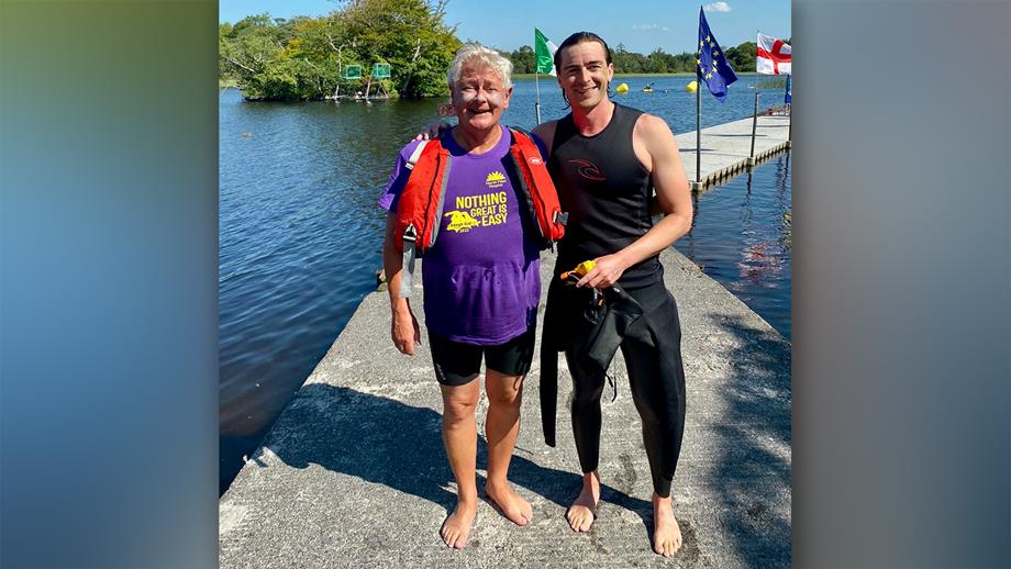 A man in a black wetsuit pulled down around his waist stands on a pontoon in a lake, with his arm around an older man, wearing a purple T-shirt. It’s a sunny day and they’re both smiling.