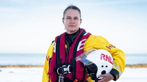 A female lifeboat crew member stands next to a rocky shore wearing inshore lifeboat kit and holding a helmet in her arms.