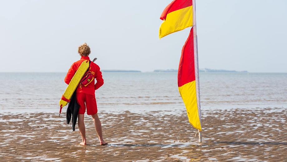 A male lifeguard is stood on the beach facing the sea with a red and yellow flag next to him.