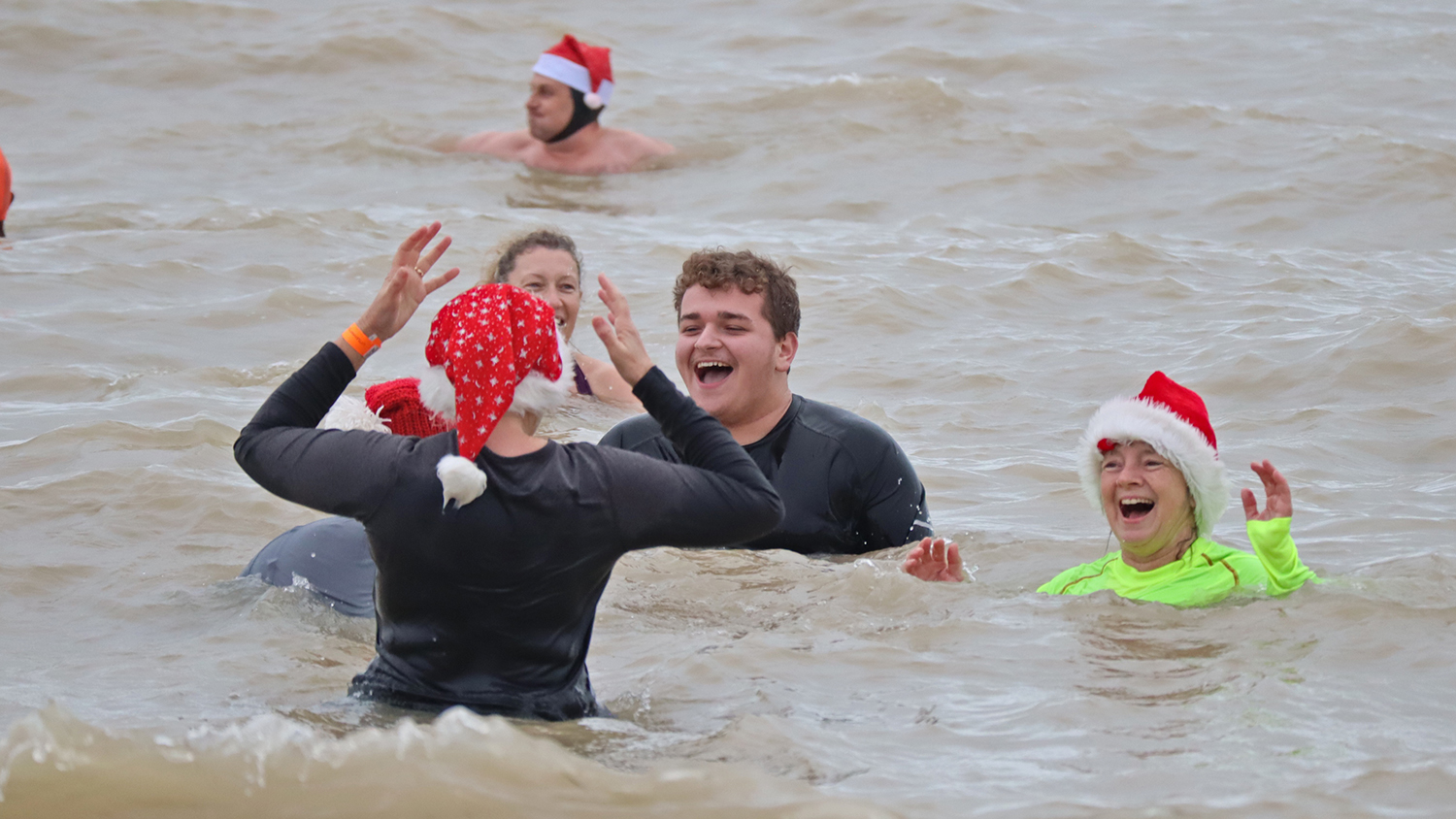 A group of people taking part in a cold water dip at Littlehampton. It’s December, so the swimmers are wearing Santa hats and many of them are wearing wetsuits. 