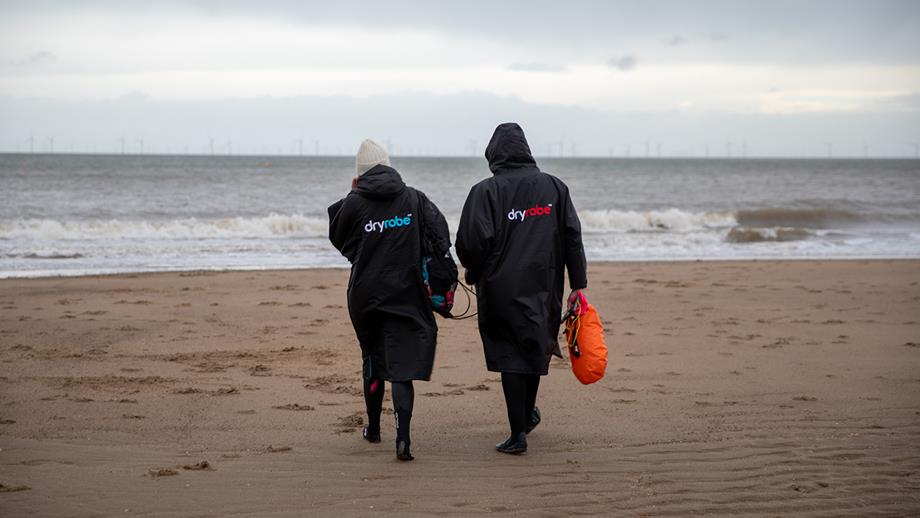 Two people walking towards the sea, wearing long dryrobes, hats, and carrying tow floats,  Caption: Wrap up warm with big coats or a dry robe