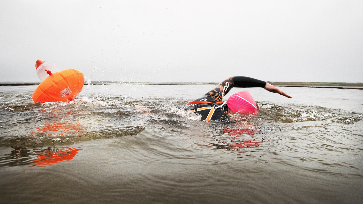A swimmer wearing a bright pink cap glides through the water, with an orange inflatable tow float bobbing behind her