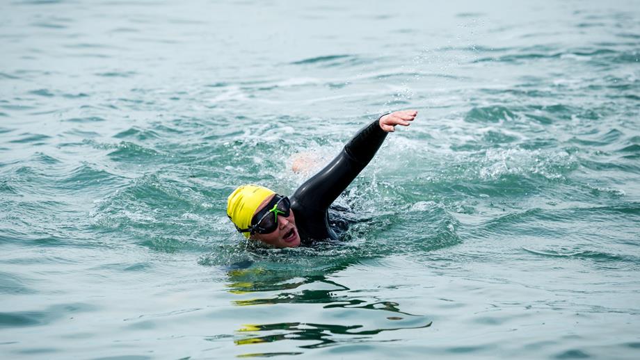 A person is swimming in open water and is wearing a swimming hat, goggles and wetsuit