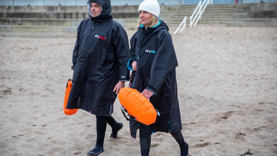 RNLI Water Safety Lead Nick Ayers and Louise Minchin walk down a sandy beach wearing Dryrobes. A weatherproof changing robe can help reduce afterdrop following open water swimming.