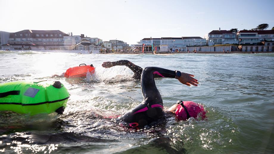 Two swimmers cut through the water in front of a beach. A flutter kick keeps your legs near the surface and conserves energy when open water swimming