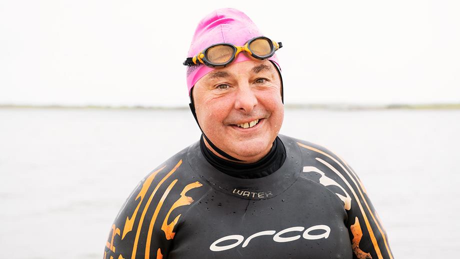 A smiley open water swimmer in front of the sea, wearing a wetsuit and brightly coloured swimming hat