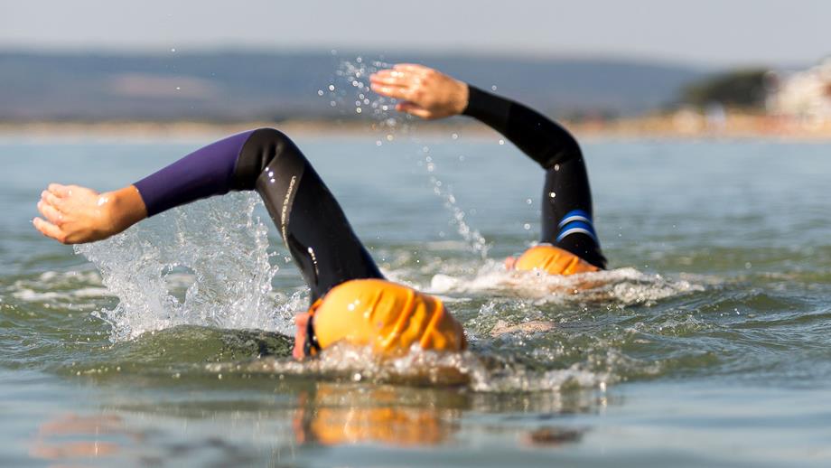 Two people are swimming in open water and are wearing wetsuits and swimming hats