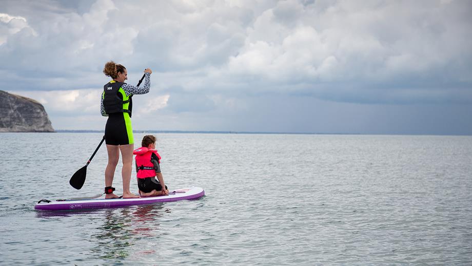 A woman paddles out to sea, with a child sitting on the front of her board. They are both wearing lifejackets.