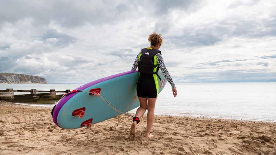 A woman wearing a wetsuit walks into the sea with a blue bodyboard, with a leash attached to her ankle