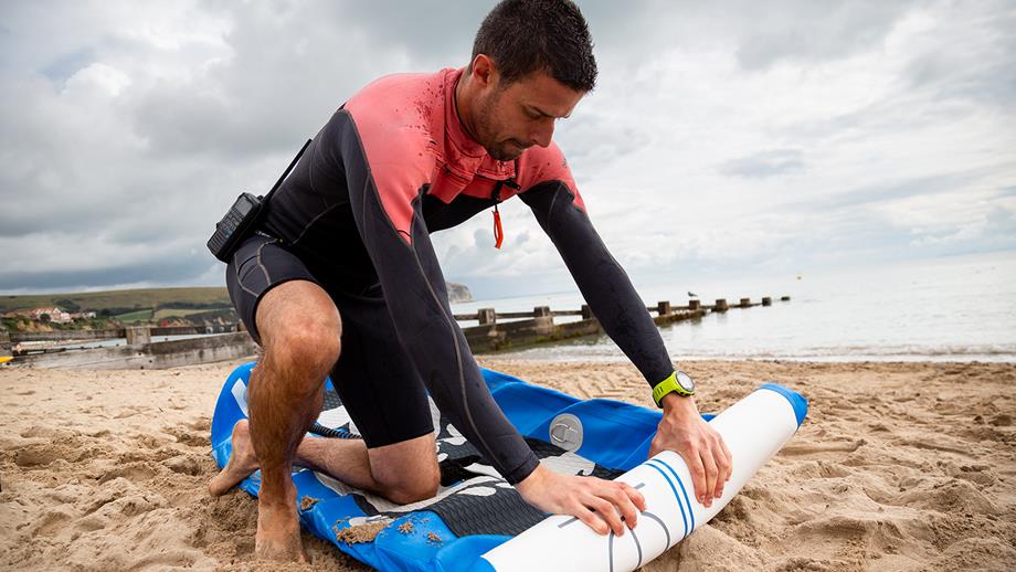 A man in a red and black wetsuit rolls up a deflated paddleboard