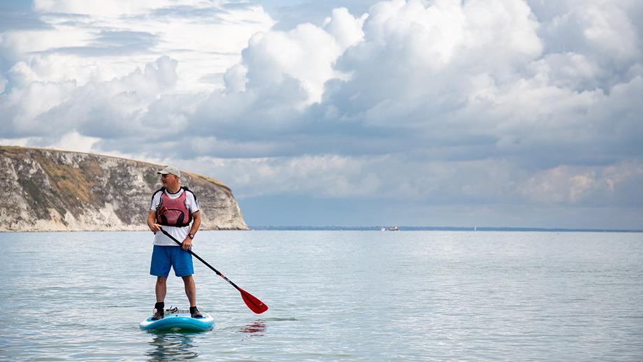 A man wearing a T-shirt, shorts and a buoyancy aid stands on a bright blue paddleboard, looking ahead
